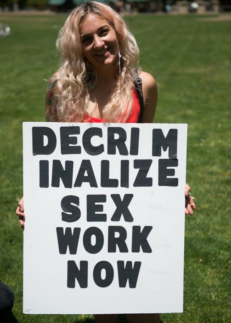 person smiling and holding a sign that says, decriminalize sex work now